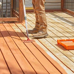 Person applying stain to a wooden deck with a paint roller.