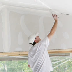 A person in white applying drywall compound to a ceiling indoors.
